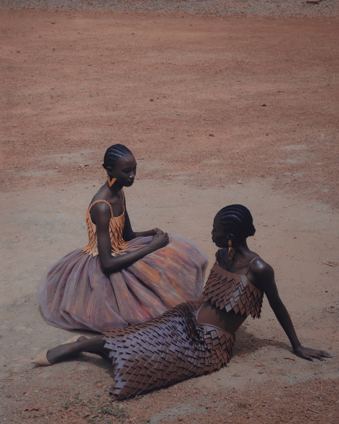 Two African models pose in earthy-toned dresses, one seated with a layered bodice and full skirt, the other reclining in a matching two-piece.