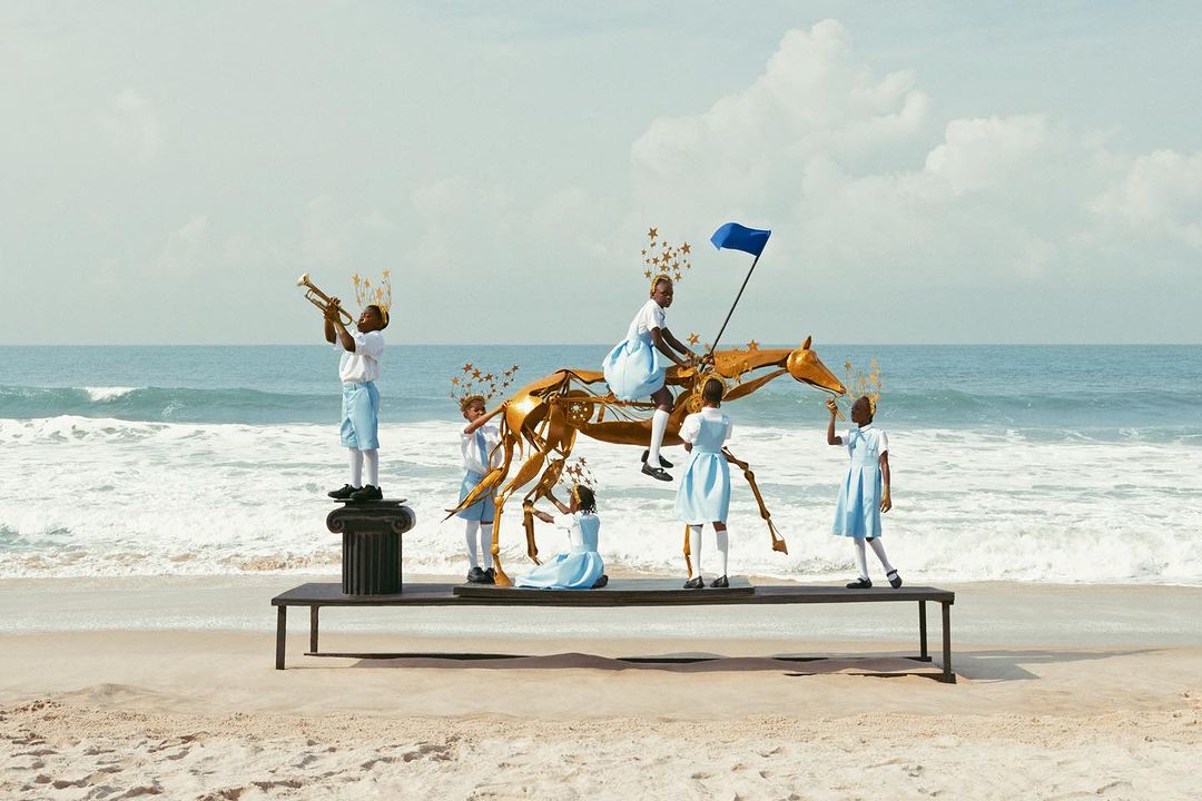 Group of children in school uniforms are posed with a gold sculpture of a horse on a table at a beach setting on a sunny day.