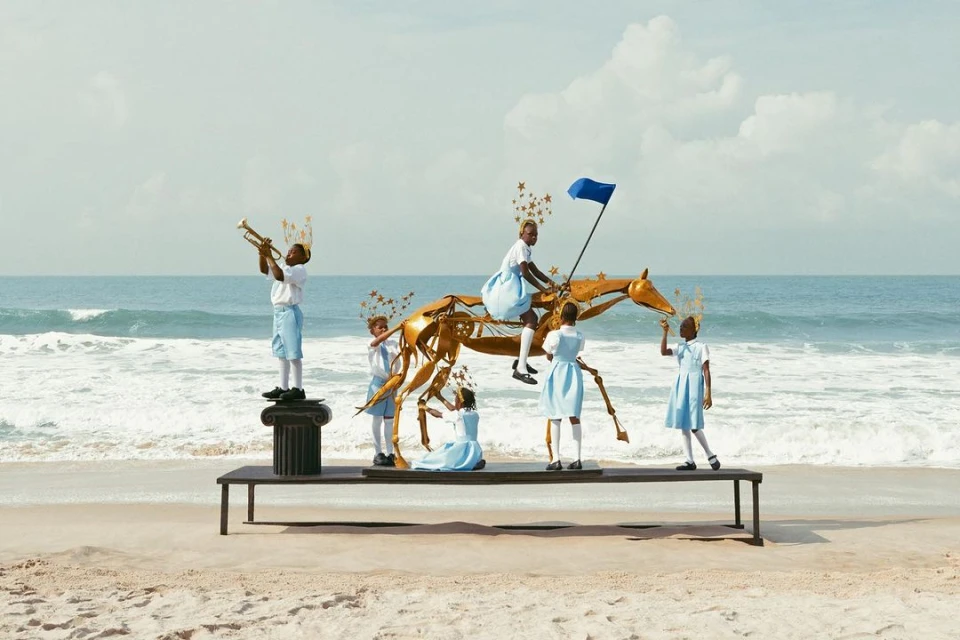 Group of children in school uniforms are posed with a gold sculpture of a horse on a table at a beach setting on a sunny day.