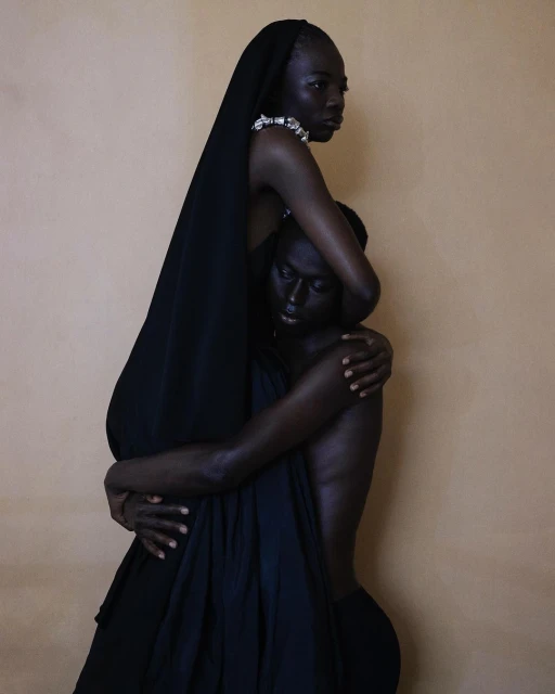 Striking portrait of two Black adult females with dark skin, embracing in a minimalist studio setting with draped black cloth and jewelry.