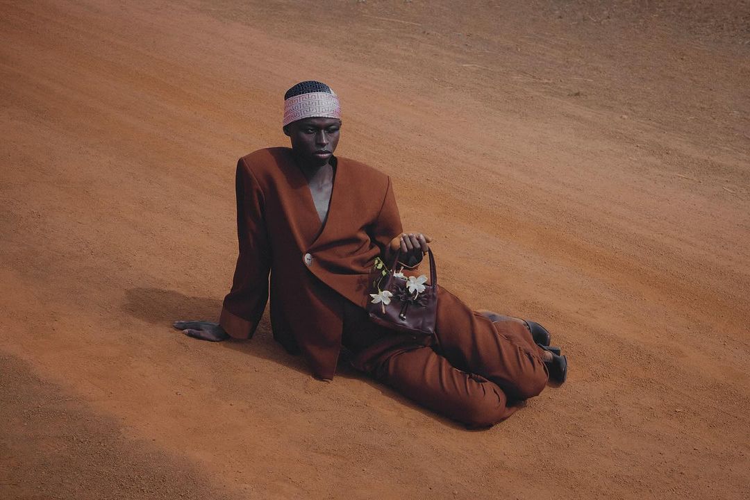 Model wearing a structured brown suit and patterned headband, sitting on a red dirt road while holding a small leather handbag with white flowers.
