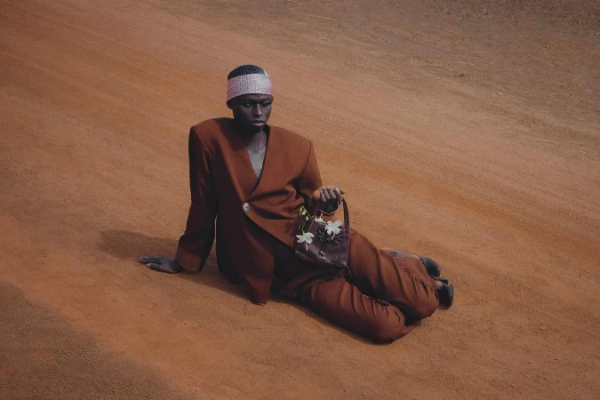 Model wearing a structured brown suit and patterned headband, sitting on a red dirt road while holding a small leather handbag with white flowers.