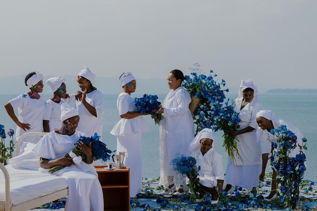 A group of nine Black women in white dresses gather with blue roses near a lake on a sunny day in a stylized, artistic outdoor scene.