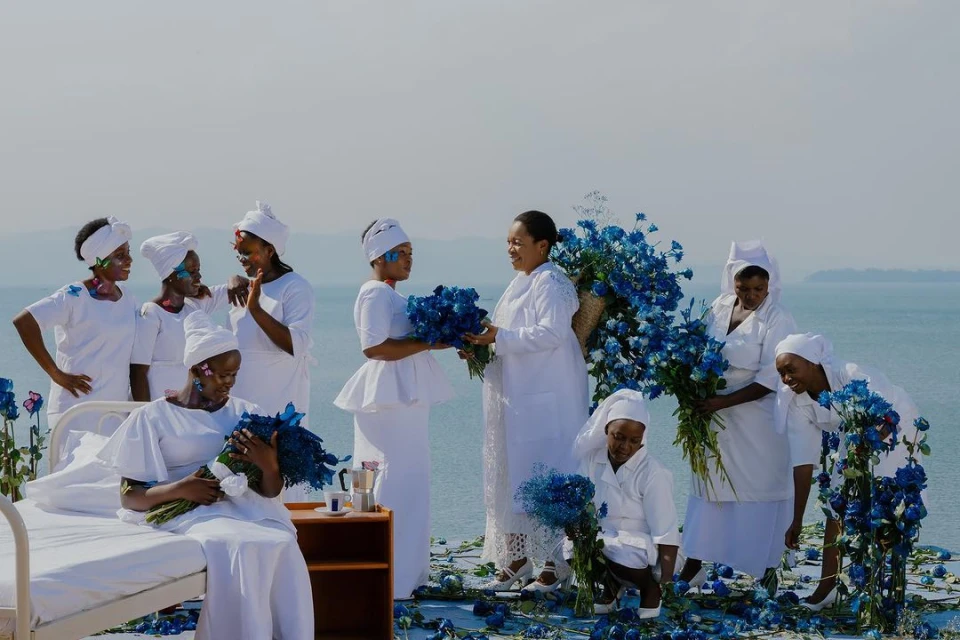 A group of nine Black women in white dresses gather with blue roses near a lake on a sunny day in a stylized, artistic outdoor scene.
