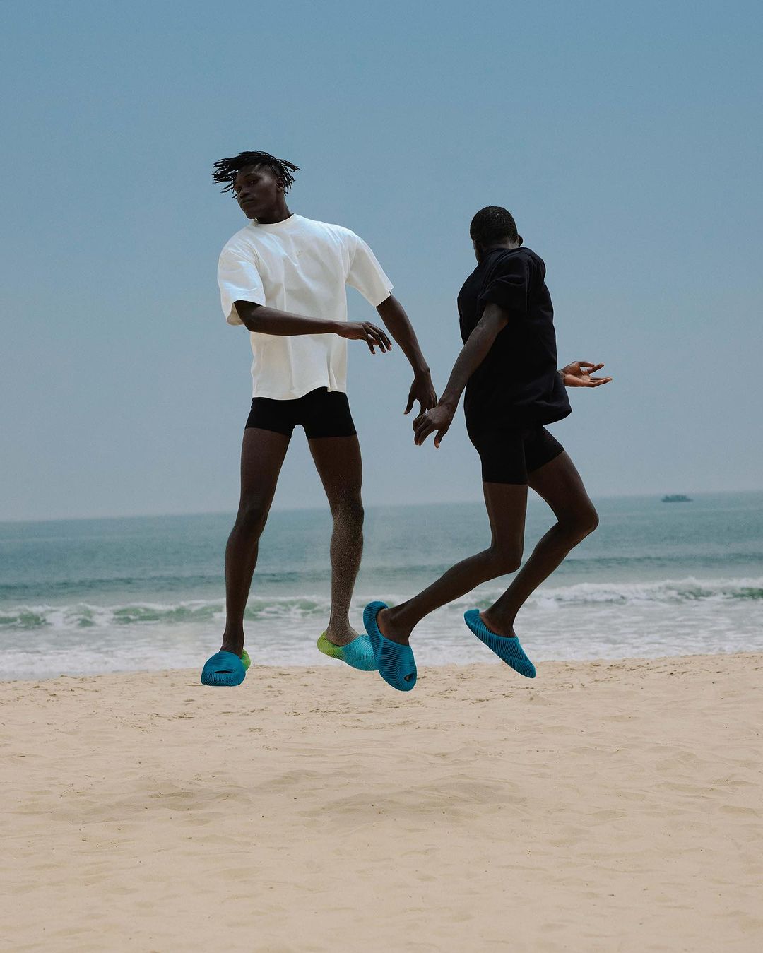 Two men leap on a sandy beach near the ocean. One wears a white t-shirt and blue slippers, the other wears black t-shirt with blue slippers.