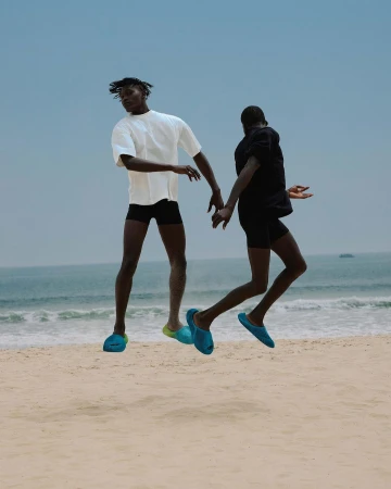 Two men leap on a sandy beach near the ocean. One wears a white t-shirt and blue slippers, the other wears black t-shirt with blue slippers.