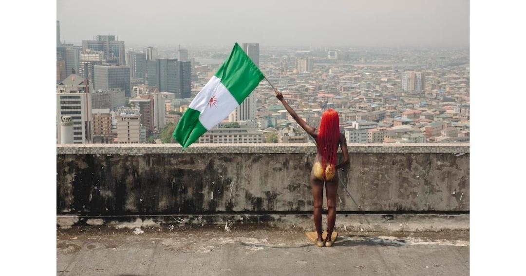 Adult woman with orange hair stands on a rooftop holding a Nigerian flag, cityscape in background, medium shot, outdoor setting.