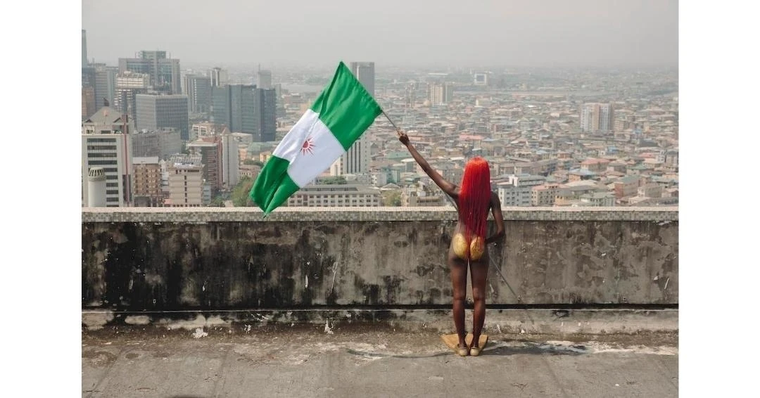 Adult woman with orange hair stands on a rooftop holding a Nigerian flag, cityscape in background, medium shot, outdoor setting.