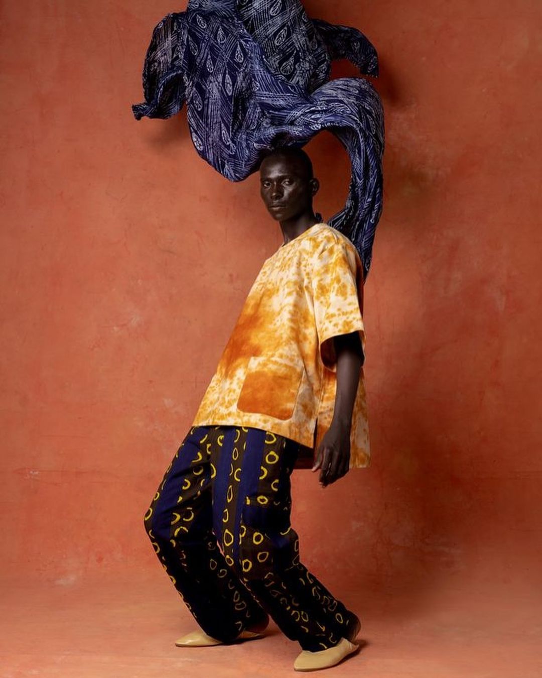 Dark-skinned model strikes a pose in a studio wearing a yellow tie-dye shirt and patterned pants, with a blue textile art piece behind.