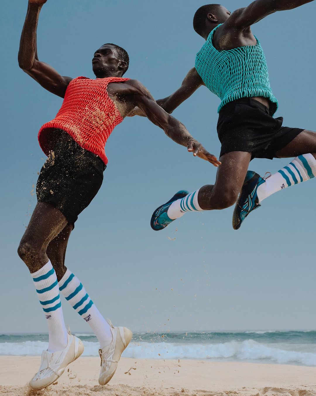Two African male athletes leap on a beach under a pale blue sky; one wears red, the other teal, both with striped socks and athletic shoes.