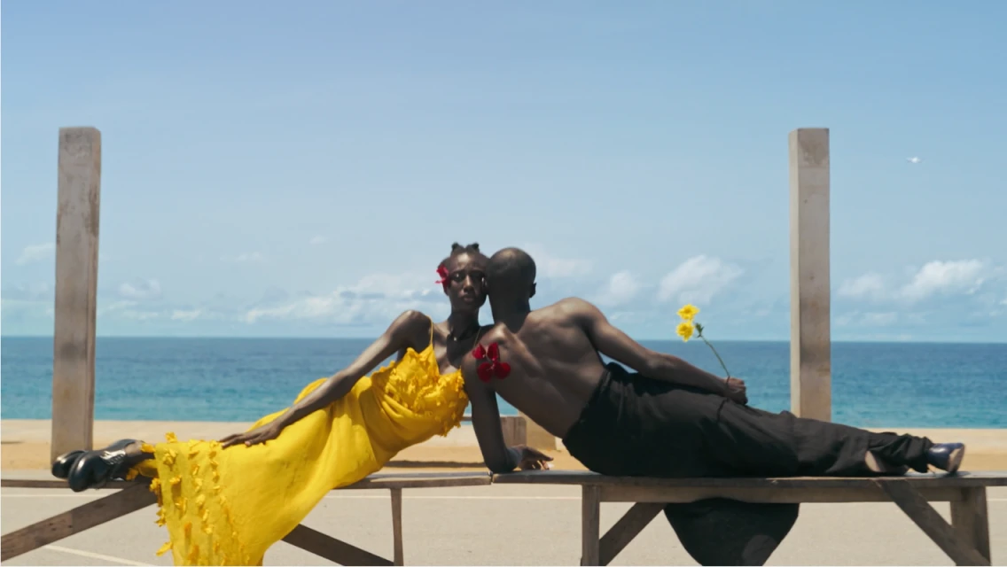 Two models on a wooden bench near a seaside. A Black woman in a yellow dress and a Black man with bare chest hold flowers.