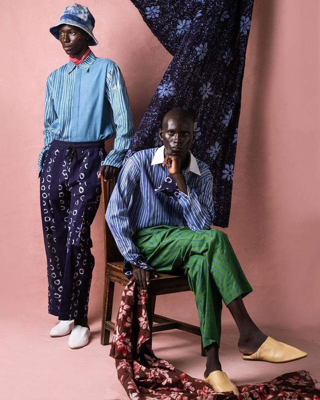 Two dark-skinned male fashion models pose against a pink backdrop, showcasing outfits with blue and green color palettes and patterned textiles.