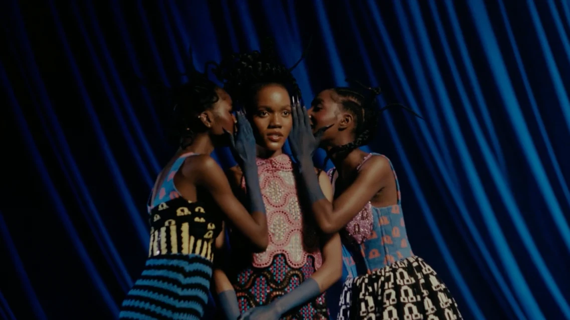 Three women in colourful, patterned dresses pose against a draped blue background. The central model is flanked by two others whispering.