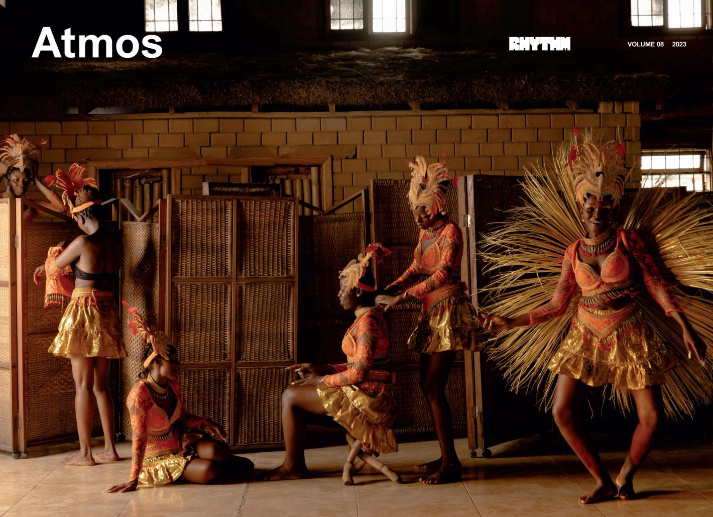 Several African women in traditional costumes with feathered headdresses pose indoors against a brick wall and woven partitions.