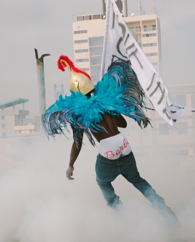 Person in blue feathers, gold helmet, and 'Barbie' briefs against city backdrop with smoke, holding a flag with writing.