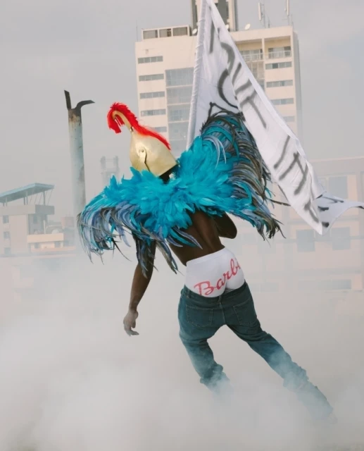 Person in blue feathers, gold helmet, and 'Barbie' briefs against city backdrop with smoke, holding a flag with writing.