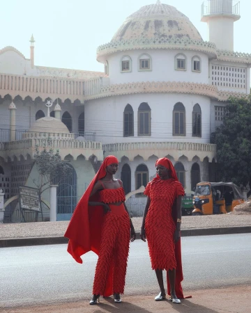 Two adult women in red outfits stand on a street near a white building with domes. A yellow vehicle is visible in the background.