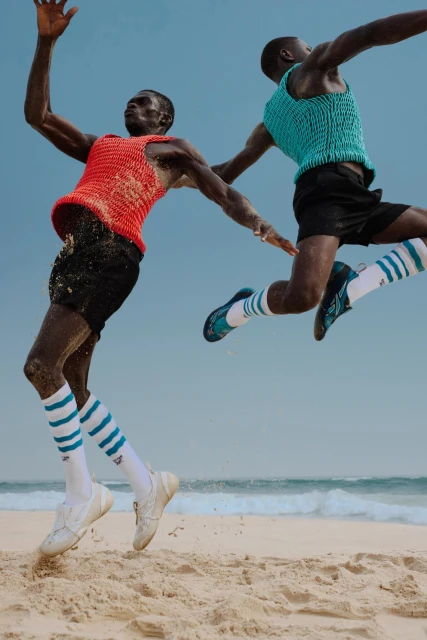 Two Black male athletes leap on a sandy beach under a clear sky, wearing colorful tank tops, black shorts, striped socks, and sneakers near the ocean.