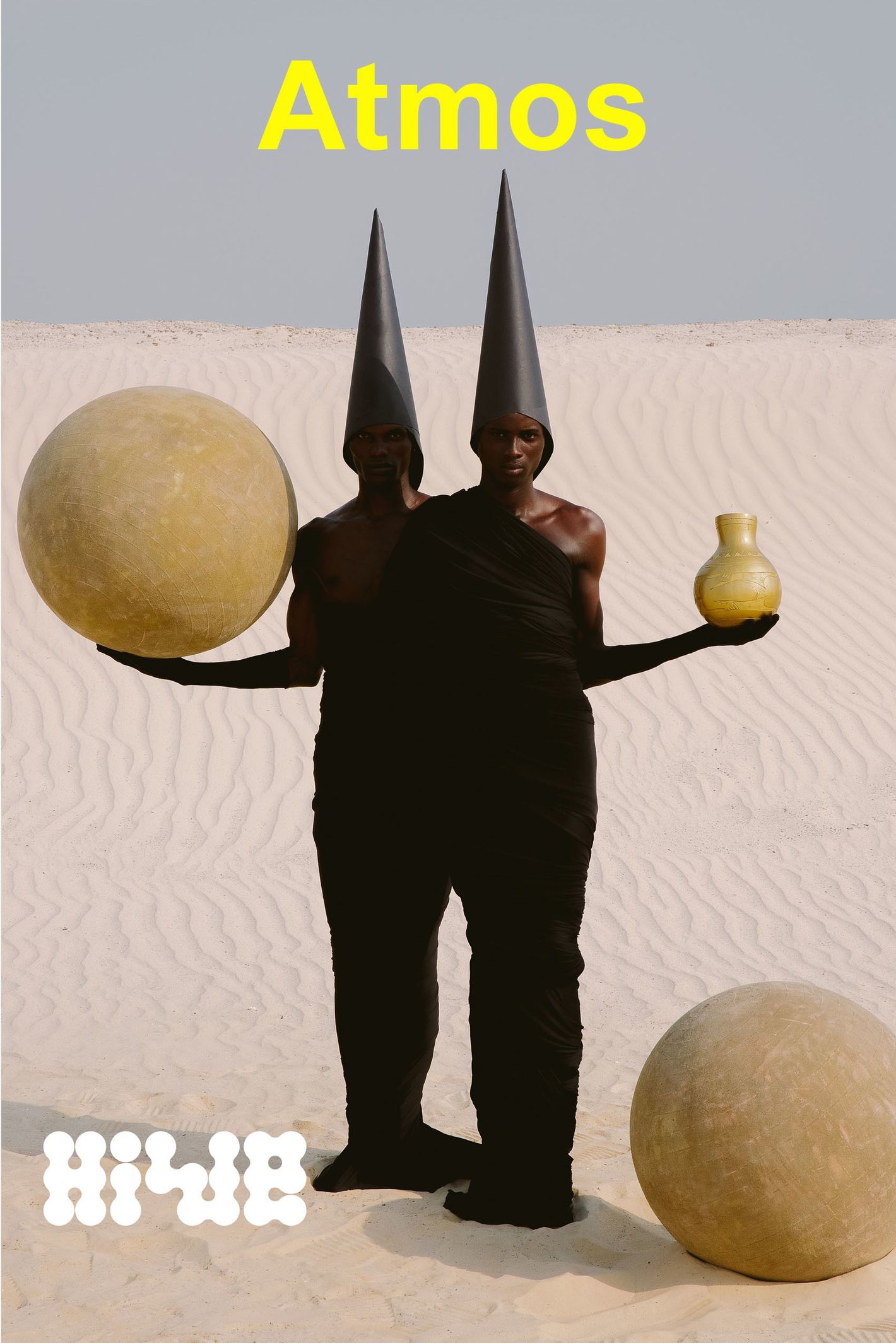 Two people in conical hats and black robes stand in a desert landscape holding an orb and a vase in this surreal outdoor scene.