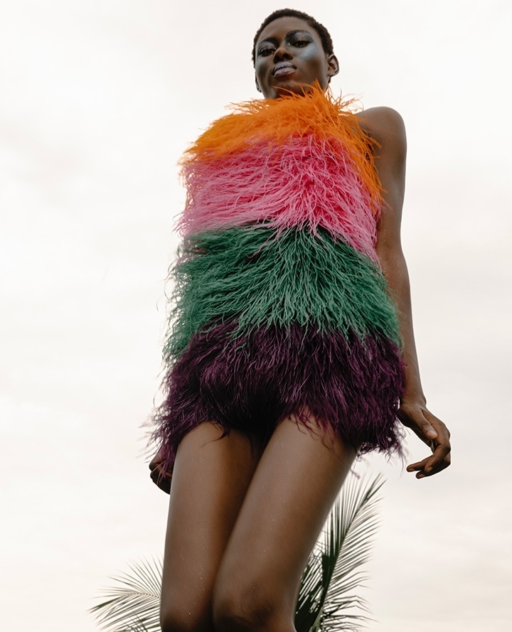 Low angle studio shot of an African woman posing in a colorful feather dress against a bright sky, evoking high fashion and nature.