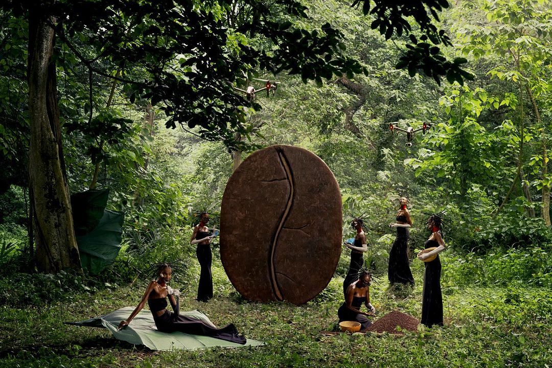 Conceptual image shows models in black dresses, a giant coffee bean sculpture, and drones in a lush green forest setting, artistic composition.