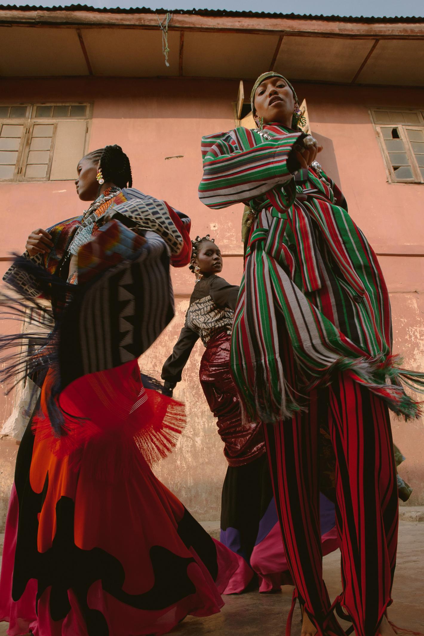 Three women pose outdoors, showcasing vividly patterned, colorful dresses. One woman is elevated on stilts, captured in a wide shot.
