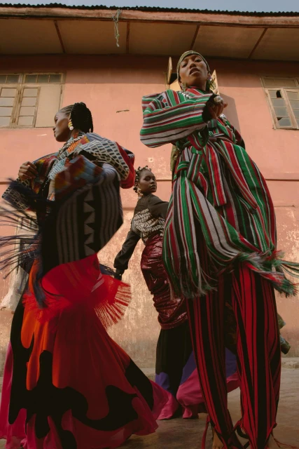 Three women pose outdoors, showcasing vividly patterned, colorful dresses. One woman is elevated on stilts, captured in a wide shot.