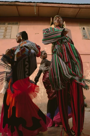 Three women pose outdoors, showcasing vividly patterned, colorful dresses. One woman is elevated on stilts, captured in a wide shot.