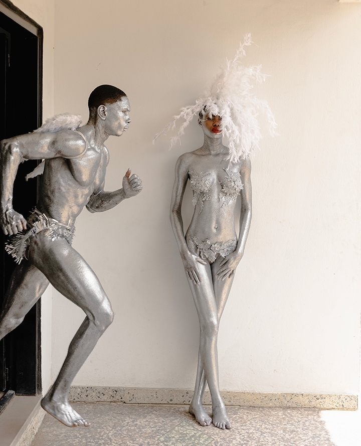 Two adults in silver body paint, one in motion wearing small wings, the other standing still with a feathered headpiece, indoors.