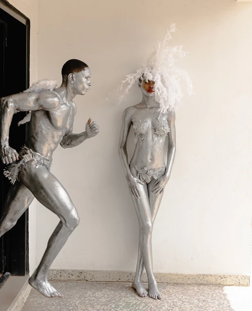 Two adults in silver body paint, one in motion wearing small wings, the other standing still with a feathered headpiece, indoors.