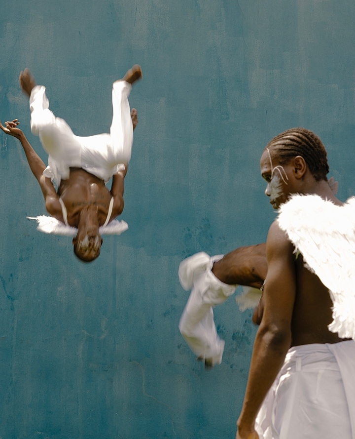 Two dark-skinned adult men perform a dance-like stunt with angel wings in an outdoor setting against a blue wall backdrop.