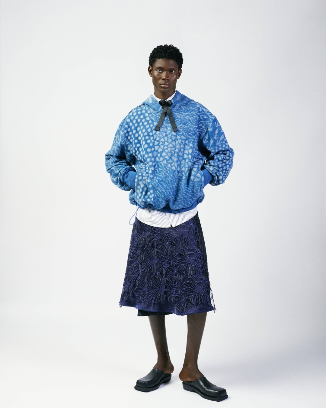 Full-length portrait of a Black male model wearing a blue patterned hoodie, navy skirt, white shirt, black bow, and black clogs on a white backdrop.