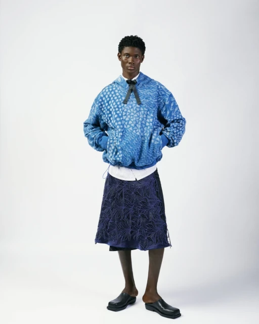 Full-length portrait of a Black male model wearing a blue patterned hoodie, navy skirt, white shirt, black bow, and black clogs on a white backdrop.