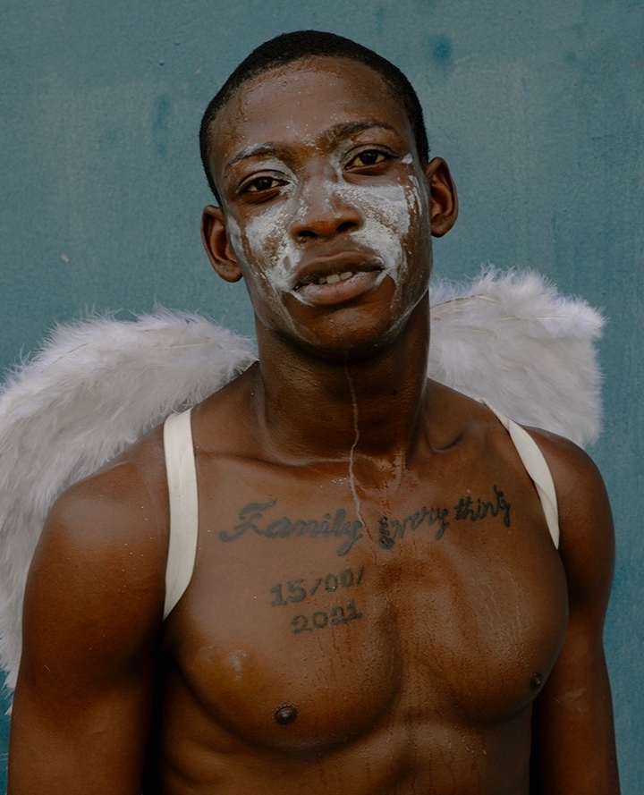 Close-up portrait of a shirtless African American man wearing angel wings and white face paint against a blue wall background.
