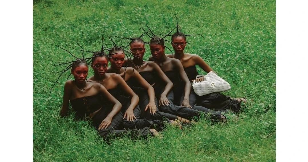 A group of six Black women are sitting in a grassy field, wearing black tube tops and dark pants, with a white purse.