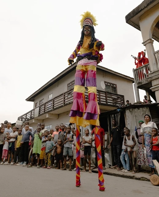 Stilt walker in colorful costume performs on street as crowd watches, houses in background.