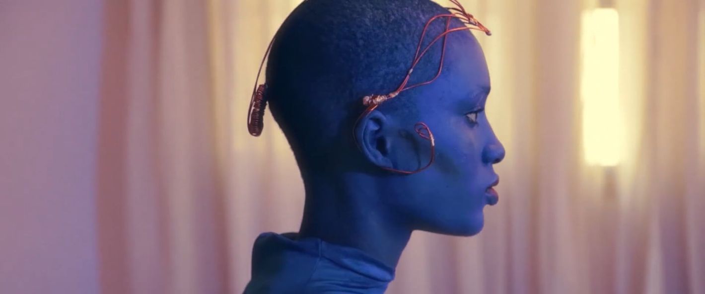Side view of a blue-skinned person with decorative wire headdress and turtleneck top, against a softly lit, blurry neutral backdrop.