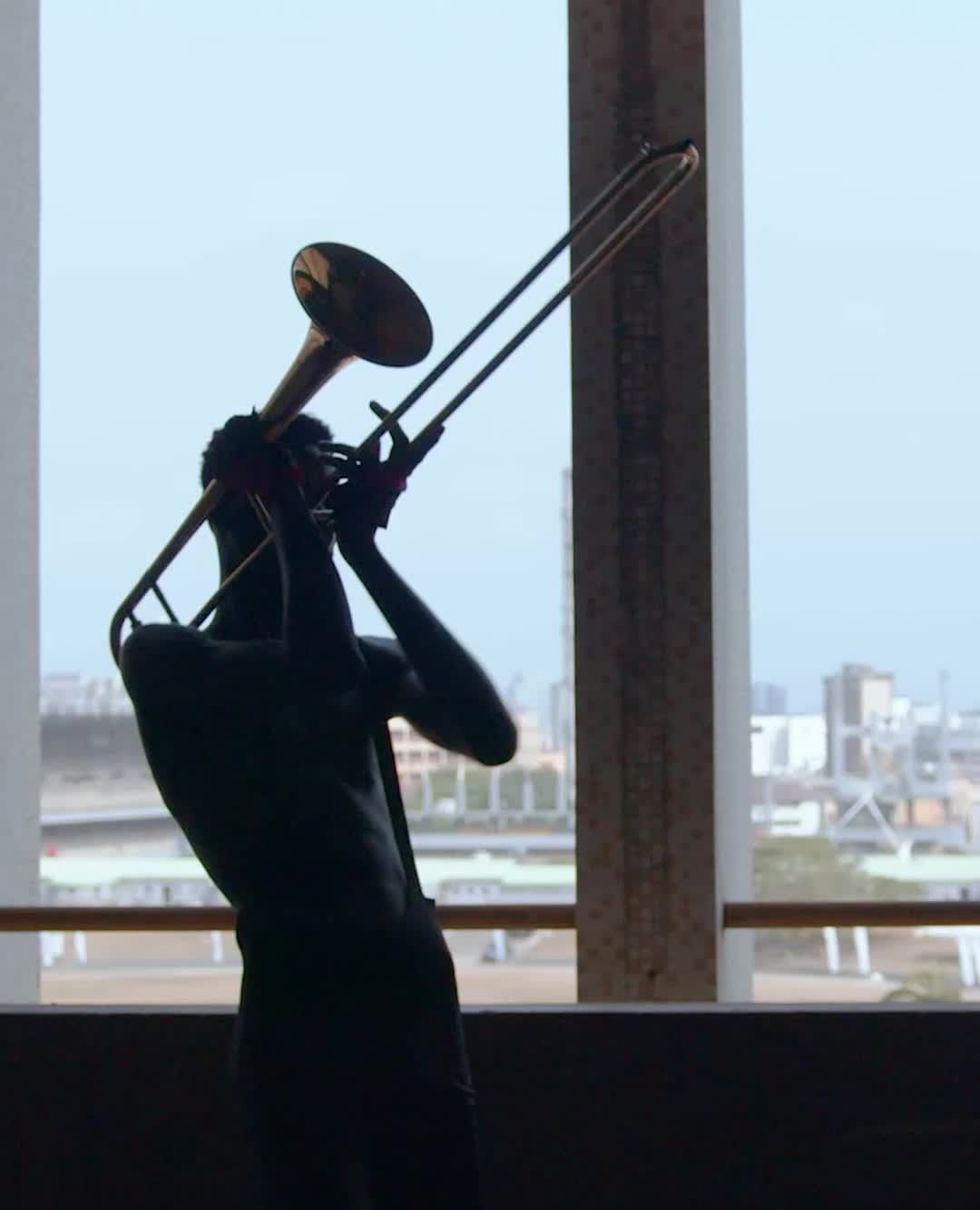 Silhouette of a person playing the trombone in a modern indoor setting with a city visible through a window.