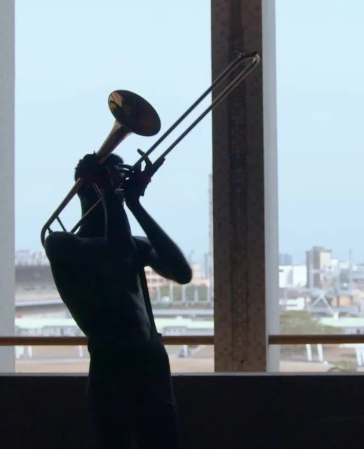 Silhouette of a person playing the trombone in a modern indoor setting with a city visible through a window.