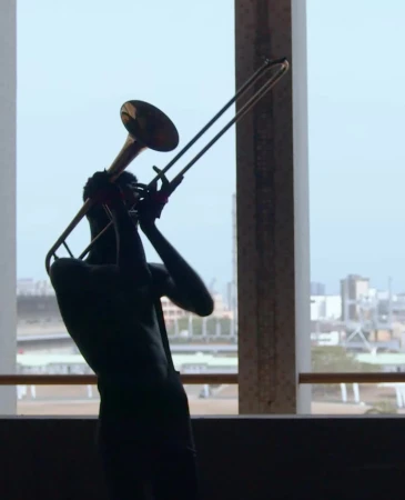 Silhouette of a person playing the trombone in a modern indoor setting with a city visible through a window.