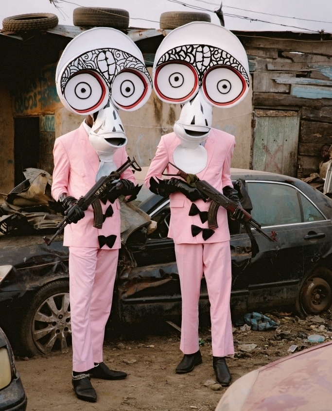 Two people in costumes stand in a dilapidated outdoor environment holding weapons, wearing masks and pink suits with bows.