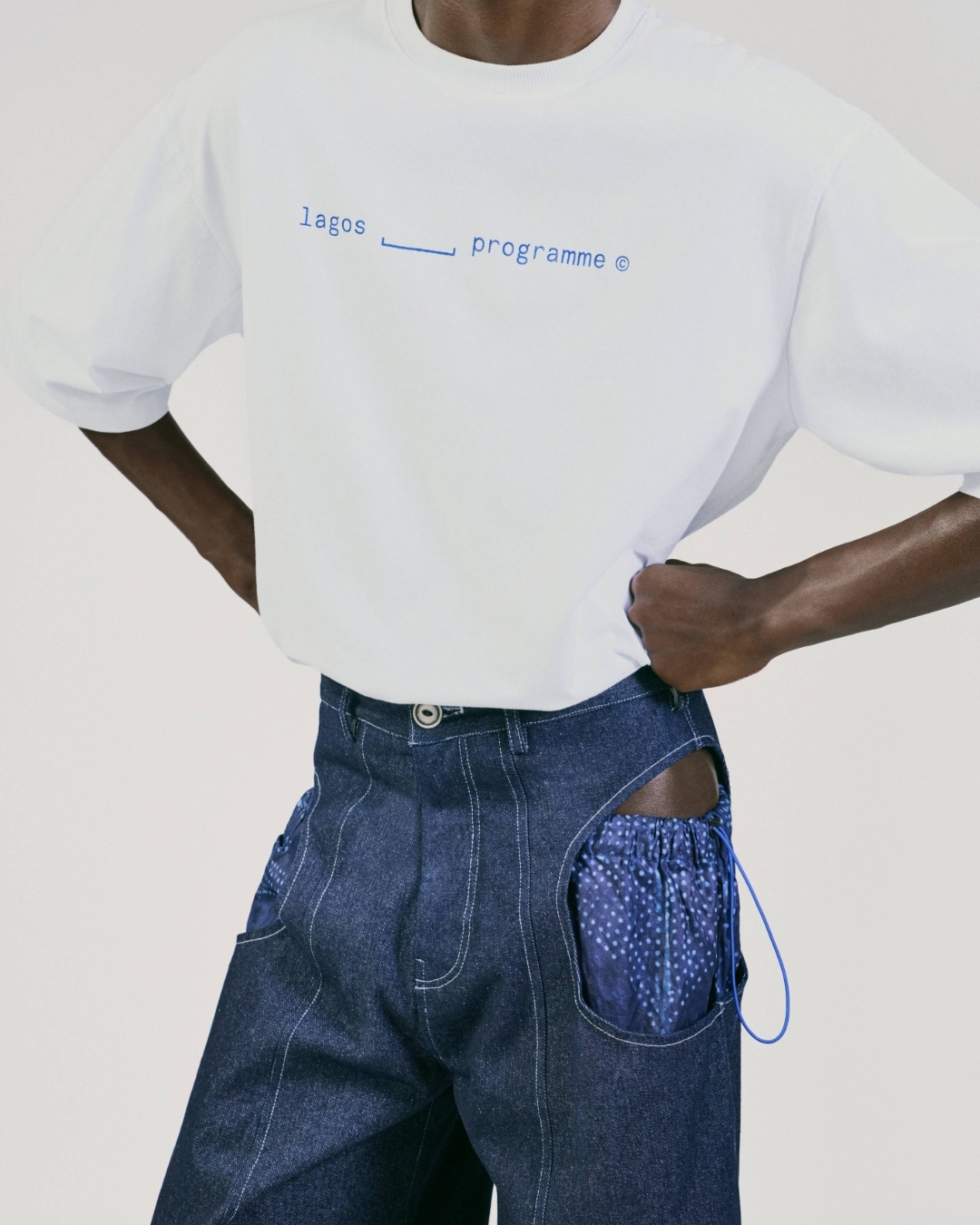 Fashion shot: a dark-skinned person poses in a white t-shirt with blue text and patchwork denim pants against a neutral backdrop.
