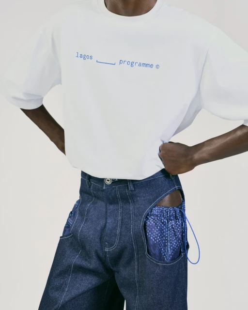 Fashion shot: a dark-skinned person poses in a white t-shirt with blue text and patchwork denim pants against a neutral backdrop.