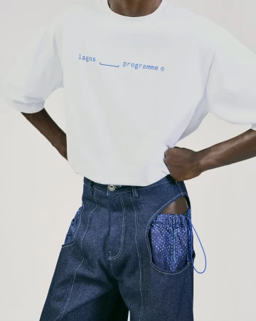 Fashion shot: a dark-skinned person poses in a white t-shirt with blue text and patchwork denim pants against a neutral backdrop.
