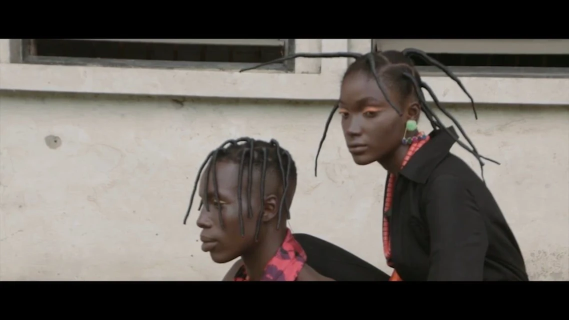 Two dark-skinned people with braids posing outdoors in front of a textured wall; both are wearing dark tops and beaded necklaces.