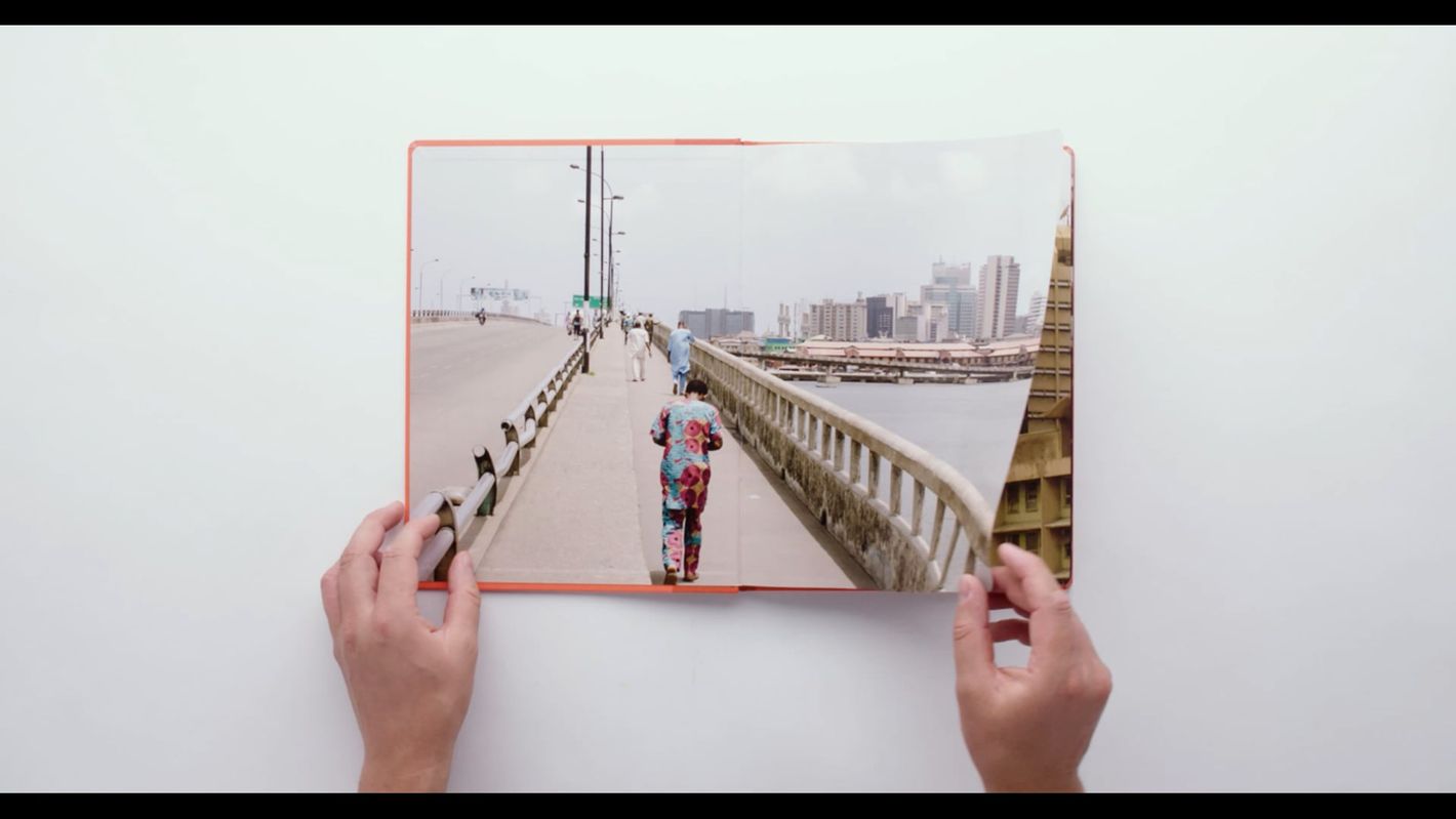 Person in patterned clothing walks along a bridge with cityscape in the distance, displayed in an open book held by hands.