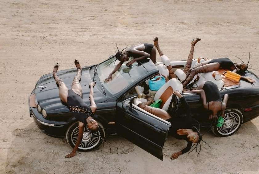 Editorial shot of a group of women contorted on and around a black convertible car in a desert landscape, wearing black bodysuits and patterned tights.