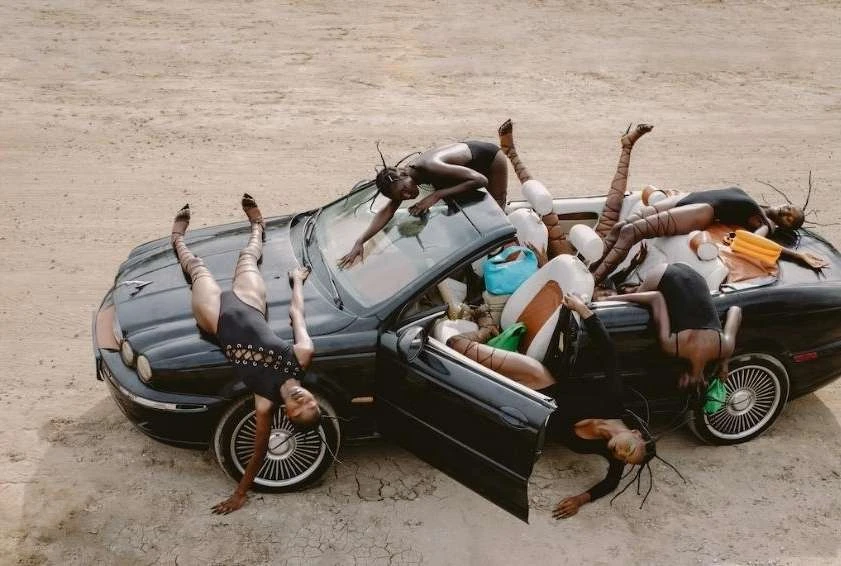 Editorial shot of a group of women contorted on and around a black convertible car in a desert landscape, wearing black bodysuits and patterned tights.