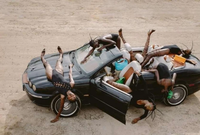 Editorial shot of a group of women contorted on and around a black convertible car in a desert landscape, wearing black bodysuits and patterned tights.