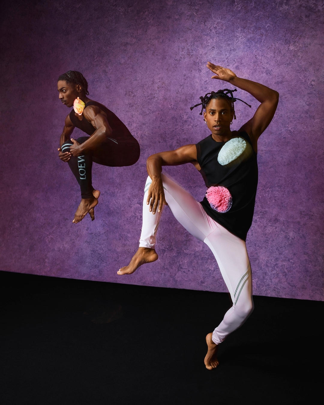Two barefoot male dancers wearing athletic clothing with colorful pom poms pose against a purple backdrop in a studio setting.